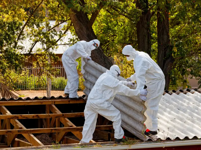 Workers in white protective suits remove corrugated roofing from a damaged roof.